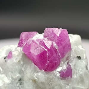 以下が含まれることがあります： 鮮やかなファセットルビーの結晶のクラスターのクローズアップ。濃いピンクがかった赤色の宝石は、白く質感のある岩を背景に配置されています。結晶は鋭いエッジと研磨された外観を示しています。