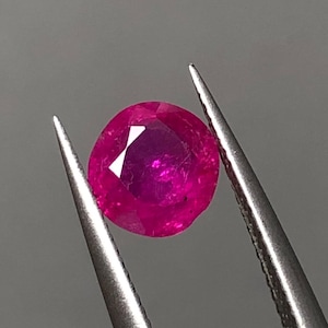 May include: A round, faceted ruby gemstone held by silver tweezers. The ruby is a vibrant, deep pinkish-red color, with visible internal facets and a polished surface. The background is a neutral gray.