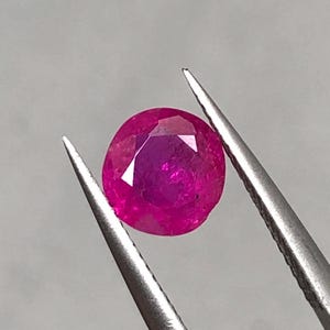 May include: A faceted, round, deep red ruby gemstone is held by a pair of silver tweezers. The ruby has a polished surface and a vibrant, saturated color. The gemstone is set against a neutral gray background.