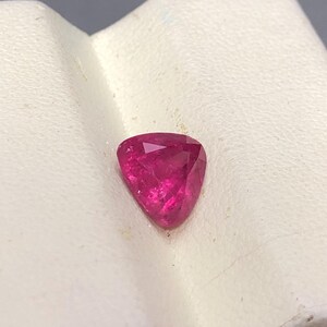 May include: A vibrant, deep pink gemstone cut into a triangular shape with multiple facets that reflect light. The stone displays a saturated, translucent color and is positioned on a textured, off-white surface with a visible diagonal crease.
