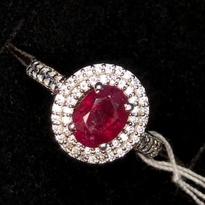 May include: A silver ring with a large, oval-cut ruby gemstone encircled by a halo of smaller, clear gemstones. The ring's band is also decorated with small gemstones, creating a sparkling effect against the dark background.