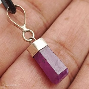 May include: A pendant featuring a rectangular, deep pink gemstone with a silver-coloured metal cap and bail. The gemstone is attached to a black cord. The pendant is held up against a neutral background.