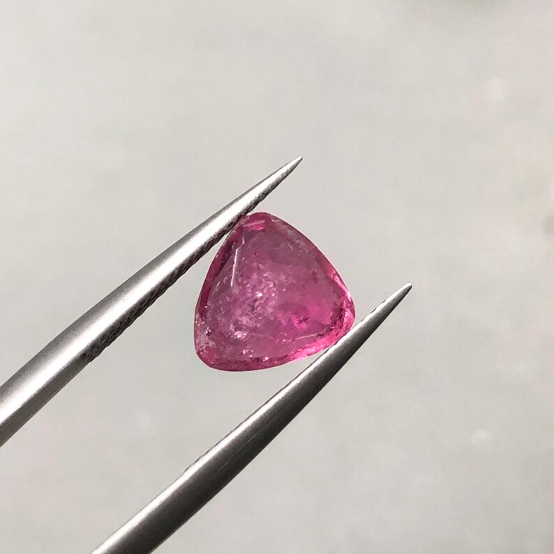 May include: A loose, triangular-cut gemstone with a vibrant, translucent hot pink hue. The stone features multiple facets that catch the light, displaying internal clarity and natural inclusions, held securely by a pair of silver-toned metal tweezers.