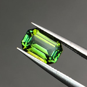 以下が含まれることがあります： エメラルドカットが施された長方形の宝石。深いフォレストグリーンから明るいライムイエローへの鮮やかなグラデーションが特徴です。銀色の金属製ピンセットで保持されており、透明感のある輝きと鋭い幾何学的なエッジがはっきりと確認できます。