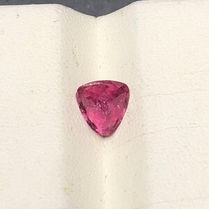 May include: A loose, faceted gemstone with a triangular shape and a vibrant, deep pink color. The stone features multiple cut surfaces that reflect light, creating a sparkling appearance across its translucent, saturated pink surface.