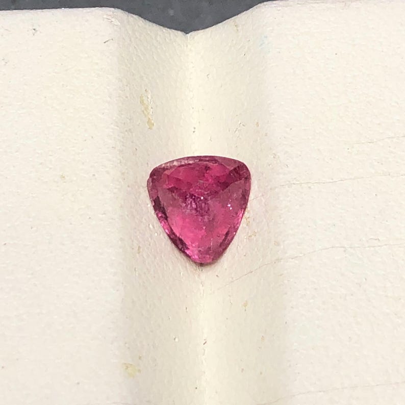 May include: A loose, faceted gemstone with a triangular shape and a vibrant, deep pink color. The stone features multiple cut surfaces that reflect light, creating a sparkling appearance across its translucent, saturated pink surface.
