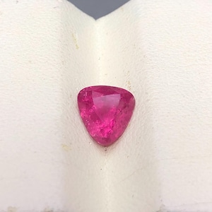 May include: A vibrant, hot pink gemstone cut into a triangular shape with multiple facets that reflect light. The stone displays a deep, saturated magenta hue throughout its surface, showcasing a clear, polished finish and sharp, defined edges.