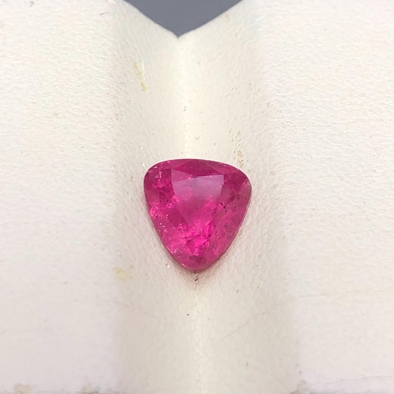 May include: A vibrant, hot pink gemstone cut into a triangular shape with multiple facets that reflect light. The stone displays a deep, saturated magenta hue throughout its surface, showcasing a clear, polished finish and sharp, defined edges.