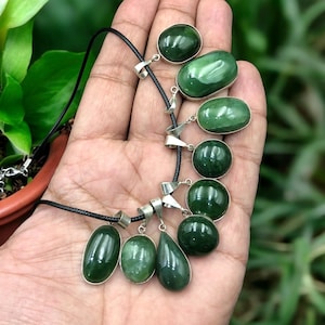 Op de afbeelding: Een ketting met meerdere donkergroene jade hangers in verschillende vormen, waaronder ovaal, rond en druppelvormig. De hangers zijn gezet in zilverkleurig metaal en geregen aan een zwart koord. De jade heeft een gepolijste, glanzende afwerking.