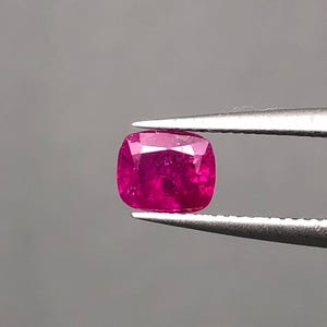 May include: A cushion-cut ruby gemstone, held by tweezers against a gray background. The ruby is a vibrant, deep pink-red color, with visible facets. The gemstone is the focal point, showcasing its clarity and brilliance.