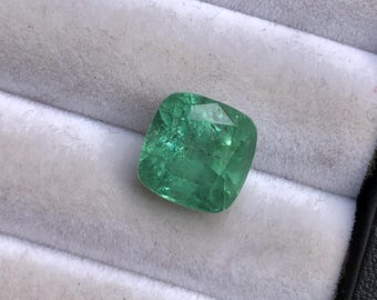 Puede incluir: Una esmeralda de talla cojín con un tono verde vibrante y una superficie facetada, que muestra inclusiones internas y reflejos de luz en toda la piedra, presentada como una pieza suelta y sin montar para joyería.