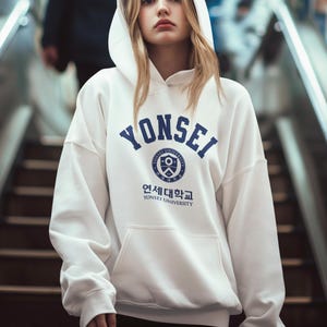 以下が含まれることがあります： 白いフード付きのスウェットシャツに、青い文字で「YONSEI」と、韓国語と英語で「YONSEI UNIVERSITY」と書かれた青いロゴが入っています。