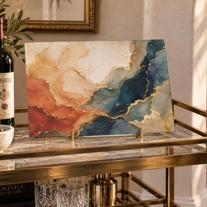 May include: A rectangular glass cutting board featuring an abstract alcohol ink design with flowing layers of deep navy blue, burnt orange, cream, and white, accented by thin, shimmering gold veins that mimic the appearance of natural stone.