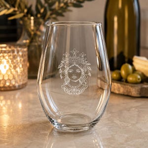 May include: A clear, stemless glass tumbler featuring a white etched graphic of a person wearing an ornate crown and jewelry. The design includes detailed facial features and decorative elements surrounding the head, centered on the glass surface.