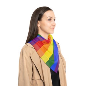 May include: A colorful rainbow-striped scarf. The scarf is draped around the neck of a person wearing a tan coat and a black shirt. The scarf features the colors of the rainbow: red, orange, yellow, green, blue, and purple.