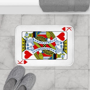 May include: A rectangular white bath mat featuring a King of Hearts playing card design. The card has a red heart suit symbol and the letter "K" in each corner. The card's design includes a blue-haired king, red and yellow details, and a white border.