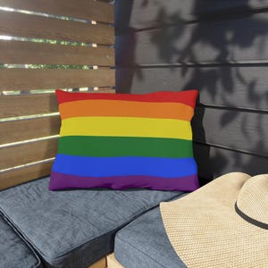 May include: Rectangular pillow with a rainbow design. The pillow features horizontal stripes in red, orange, yellow, green, blue, and purple. The pillow is placed on a blue cushion, with a straw hat visible in the corner.