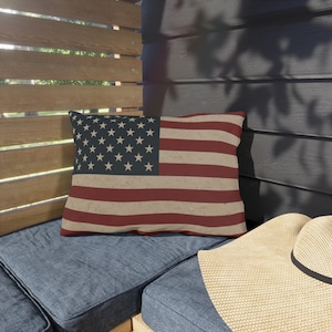 May include: A rectangular pillow with the American flag design. The pillow features a blue field with white stars and red and white stripes. The pillow is placed on a blue cushion, with a straw hat in the foreground.