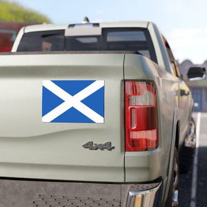 Pode incluir: Um íman retangular com a bandeira escocesa, uma cruz branca sobre fundo azul, está fixado na parte traseira de uma pick-up verde clara. O camião tem um emblema 4x4.