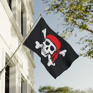 Puede incluir: Bandera negra con un diseño de calavera y huesos cruzados blancos, con un pañuelo rojo. La bandera está montada en un mástil plateado y se muestra al aire libre, con un edificio y árboles al fondo.