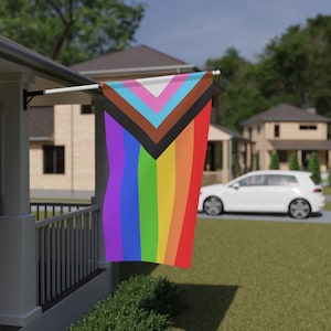 May include: A Pride flag with a progressive design, featuring a rainbow of colors and additional stripes representing inclusion. The flag is displayed outdoors, attached to a white pole on a porch, with houses and a car in the background.