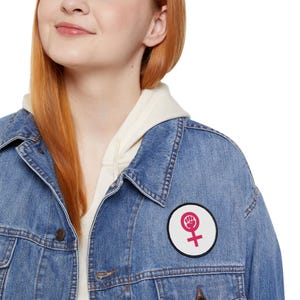 Könnte beinhalten: Ein runder Aufnäher mit schwarzem Rand, der ein pinkfarbenes weibliches Symbol und eine geballte Faust auf weißem Grund zeigt. Der Aufnäher ist auf einer blauen Jeansjacke angebracht.