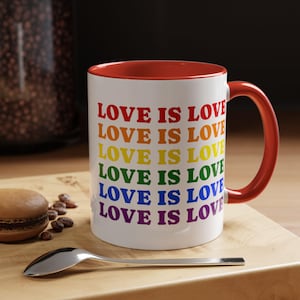 May include: A white ceramic mug with a red interior and handle. The mug features the text "LOVE IS LOVE" in rainbow colors. The mug is on a wooden surface with a spoon and a macaron.