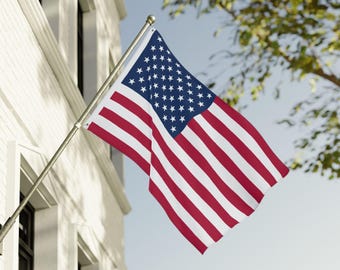 May include: American flag waving in the wind, mounted on a silver pole. The flag features red and white stripes and a blue canton with white stars. The flag is displayed outside a white building on a sunny day.