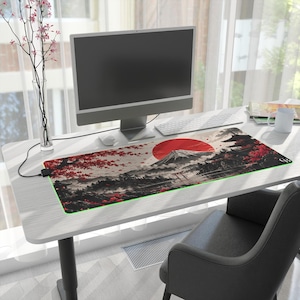 May include: A rectangular desk mat featuring a printed landscape of a mountain, a large red sun, and cherry blossom trees. The mat has a black border with integrated LED lighting and a power cable extending from the left side of the surface.