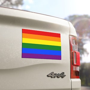 Könnte beinhalten: Ein rechteckiger Magnet mit der Regenbogenflagge, mit horizontalen Streifen in Rot, Orange, Gelb, Grün, Blau und Lila. Der Magnet ist an der Rückseite eines weißen Fahrzeugs angebracht.