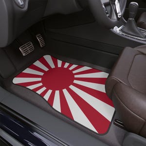 Könnte beinhalten: Eine Auto-Fußmatte mit einem roten und weißen Sonnenstrahl-Design. Die Matte ist rechteckig und für den Fußraum der Fahrerseite konzipiert. Der Innenraum des Autos ist sichtbar, einschließlich Sitz und Pedalen.
