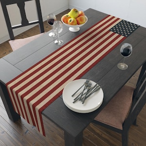 Puede incluir: Un camino de mesa con un diseño que se asemeja a la bandera estadounidense, con rayas rojas y crema y un cuadrado azul con estrellas blancas. El camino de mesa está colocado sobre una mesa de madera oscura, con platos, cubiertos y copas de vino.
