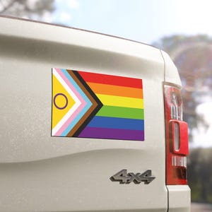 May include: Rectangular car magnet featuring the Progress Pride flag design. The flag combines the traditional rainbow flag with the transgender flag, the intersex flag, and colors representing marginalized communities of color. The magnet is affixed to a white vehicle.
