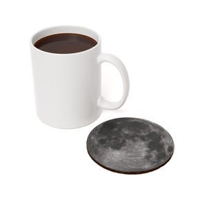May include: A white ceramic mug filled with dark liquid, likely coffee, sits next to a round coaster. The coaster features a detailed image of the moon's surface. The mug has a curved handle.