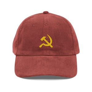 May include: A brick-red corduroy baseball cap featuring a yellow hammer and sickle emblem on the front. The cap has a curved brim and an adjustable strap at the back, suitable for various head sizes.