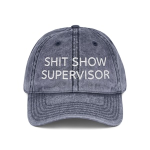 May include: A denim-colored baseball cap with the words "SHIT SHOW SUPERVISOR" embroidered in white. The cap has a curved brim and an adjustable strap at the back. The hat is a vintage style.