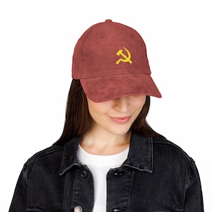 May include: A corduroy baseball cap in a reddish-brown hue, featuring a yellow embroidered hammer and sickle emblem on the front. The cap has a curved brim and a structured crown. The hat is a casual accessory.