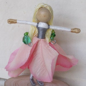 K&ouml;nnte beinhalten: Eine handgefertigte Puppe mit blonden Garnhaaren, einem rosa Stoffkleid und gr&uuml;nen Perlenakzenten. Die Puppe hat einen Holzkopf und wei&szlig;e Garnarme.