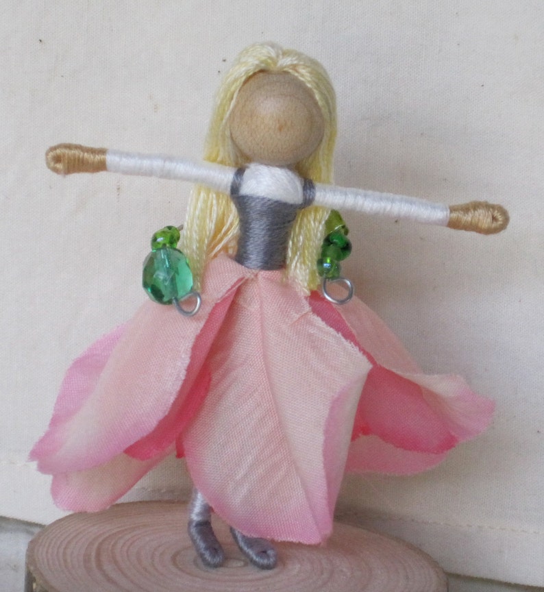 K&ouml;nnte beinhalten: Eine handgefertigte Puppe mit blonden Garnhaaren, einem rosa Stoffkleid und gr&uuml;nen Perlenakzenten. Die Puppe hat einen Holzkopf und wei&szlig;e Garnarme.