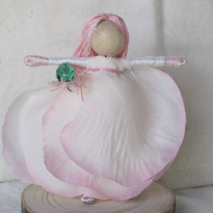 K&ouml;nnte beinhalten: Eine handgefertigte Puppe mit pinkfarbenem Garnhaar, einem wei&szlig;en Kleid mit rosa R&auml;ndern und einem gr&uuml;nen Edelsteinanh&auml;nger. Die Puppe steht auf einem Holzsockel.