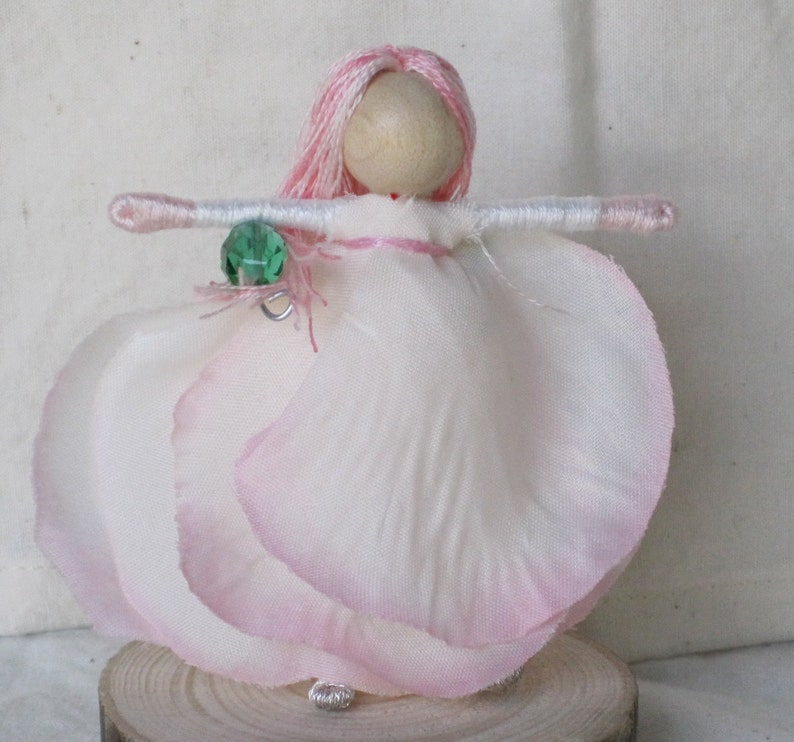 K&ouml;nnte beinhalten: Eine handgefertigte Puppe mit pinkfarbenem Garnhaar, einem wei&szlig;en Kleid mit rosa R&auml;ndern und einem gr&uuml;nen Edelsteinanh&auml;nger. Die Puppe steht auf einem Holzsockel.