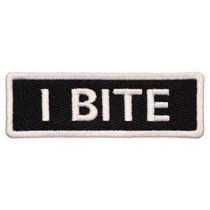 Op de afbeelding: Zwart-witte geborduurde patch met witte tekst "I BITE" op een zwarte achtergrond.