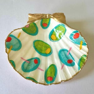 May include: A white scallop shell decorated with a pattern of green, blue, and red olives. The olives have yellow or red centers and are outlined in blue. The shell's edges and top are trimmed with gold paint.