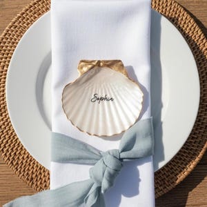 Peut inclure: Une assiette blanche avec une serviette en tissu blanc nouée par un ruban vert sauge. Un coquillage blanc aux bords peints en doré, portant le prénom Sophia écrit en noir, est posé sur la serviette. Une fourchette et un couteau dorés sont présents.