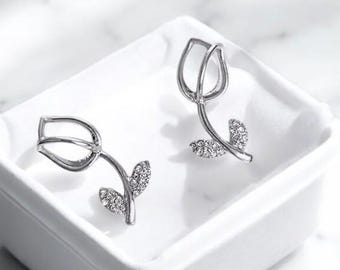 May include: A pair of silver-toned tulip earrings. Each earring features a tulip flower design with a stem and two leaves. The leaves and the base of the flower are embellished with small, sparkling stones. The earrings are displayed in a white square dish.