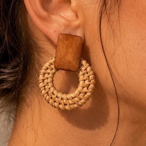 Può includere: Orecchini con un perno quadrato in legno e un cerchio in rattan intrecciato. Gli orecchini sono di colore beige naturale e hanno uno stile boho. Un accessorio elegante.