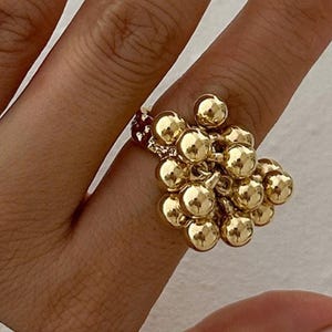 Op de afbeelding: Een goudkleurige ring met een cluster van kleine, ronde bollen. De bollen zijn zo gerangschikt dat ze een textuurrijk, opvallend ontwerp creëren. De ring wordt aan een vinger gedragen en toont de ingewikkelde details en het reflecterende oppervlak.