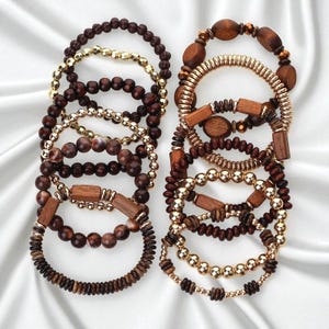 Puede incluir: Una colección de pulseras apiladas con una variedad de diseños. Las pulseras incluyen cuentas de madera, cuentas de metal color oro y detalles rectangulares de madera. La paleta de colores consiste en tonos marrones, dorados y de madera natural, creando una estética cálida y terrosa.