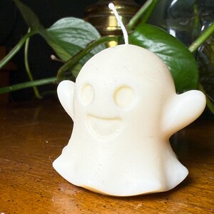 May include: A cream-coloured ghost-shaped candle with a smiling face, two eye holes, and outstretched arms. The candle sits on a wooden surface, with green foliage and a brass object in the background. A white wick is visible.