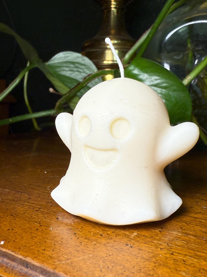 May include: A cream-coloured ghost-shaped candle with a smiling face, two eye holes, and outstretched arms. The candle sits on a wooden surface, with green foliage and a brass object in the background. A white wick is visible.